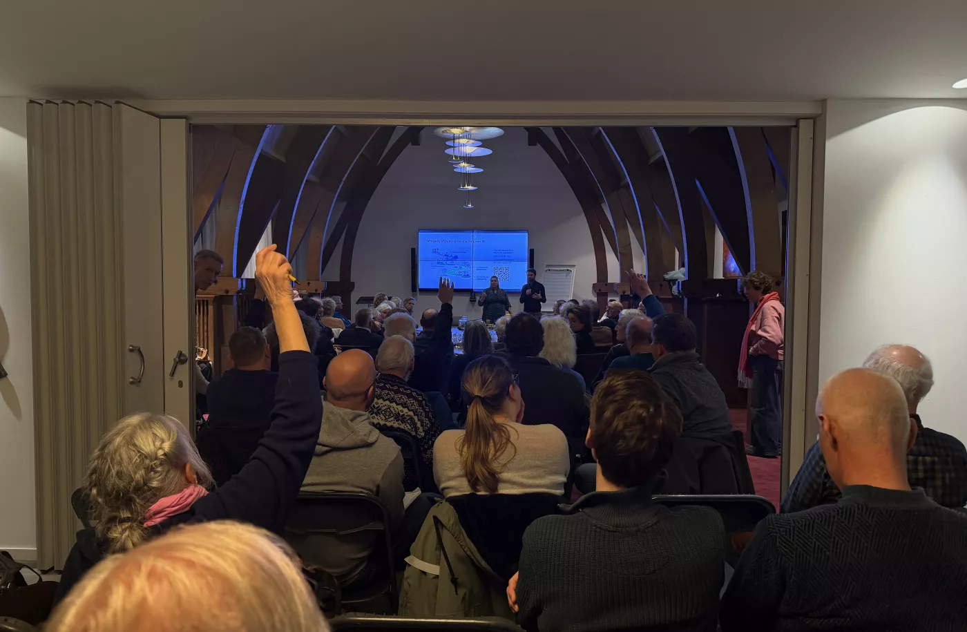 Een volle zaal met inwoners die zitten en luisteren naar een presentatie tijdens de informatiebijeenkomst voor VvE's. Voorin staat een spreker naast een groot scherm met een presentatie. Een persoon in het publiek steekt een hand op om een vraag te stellen.