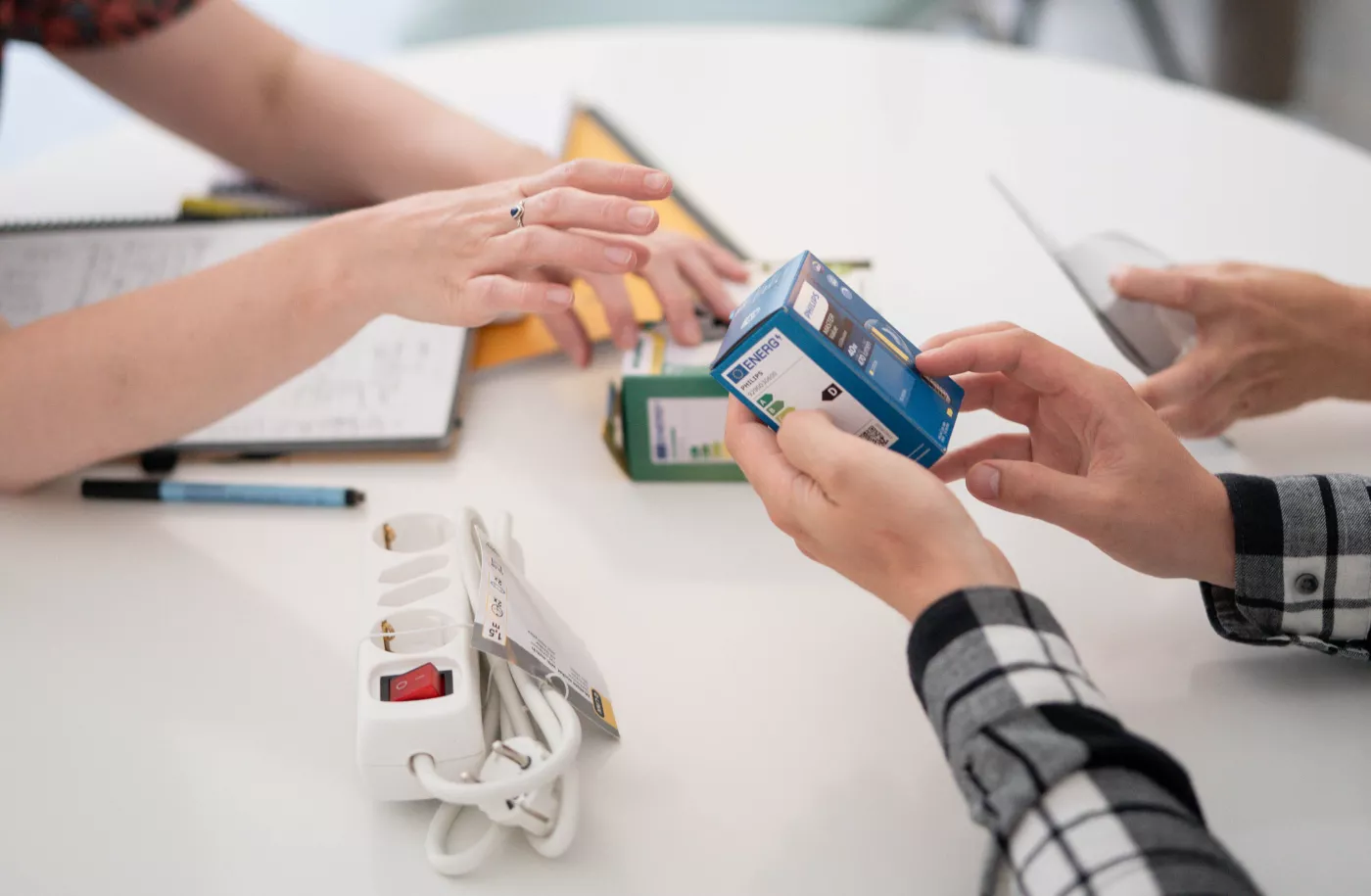 Je ziet twee paar handen die op een tafel energiebesparende producten pakken, zoals een ledlamp en een stekketblok met schakelaar 