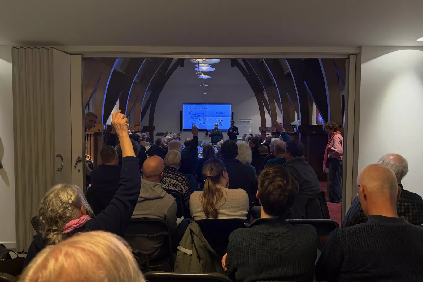 Een volle zaal met inwoners die zitten en luisteren naar een presentatie tijdens de informatiebijeenkomst voor VvE's. Voorin staat een spreker naast een groot scherm met een presentatie. Een persoon in het publiek steekt een hand op om een vraag te stellen.